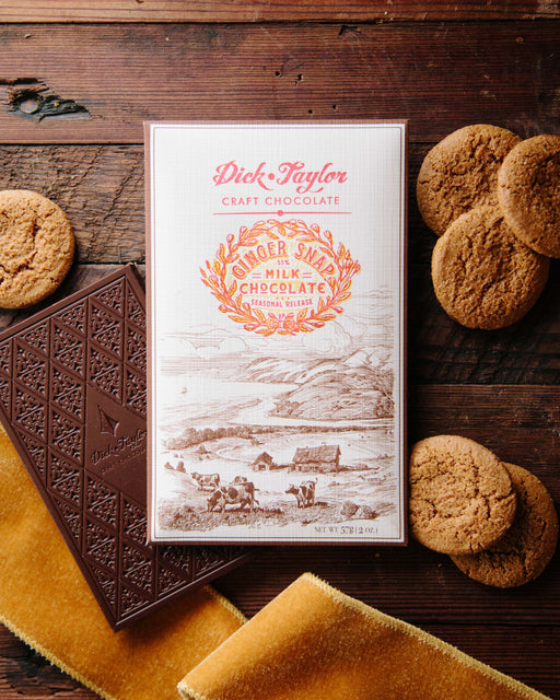 Ginger Snap Milk Chocolate bar on top of an unwrapped bar on a wood background decorated with ginger snap cookies and yellow ribbon.